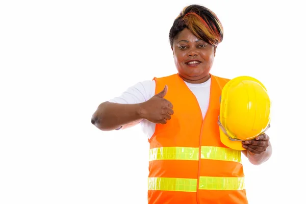 スタジオショットの幸せな脂肪黒アフリカの女性建設労働者の笑顔ながら 安全ヘルメットを保持し 親指を放棄 — ストック写真