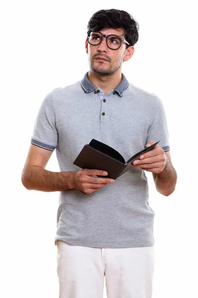 Studio shot of young Persian man holding book while thinking and — Stock Photo, Image