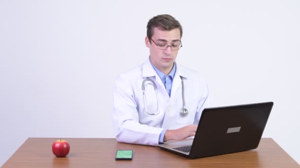 Young happy handsome man doctor video calling at work against wooden table — Stock Video
