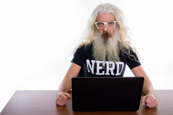 Studio shot of senior bearded nerd man using laptop on wooden ta
