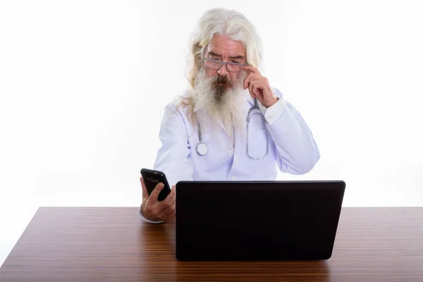Studio shot of senior bearded man doctor using mobile phone with — Stock Photo, Image