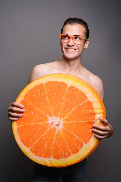 Jovem bonito homem sem camisa segurando grande fatia de fruta laranja enquanto pensa — Fotografia de Stock