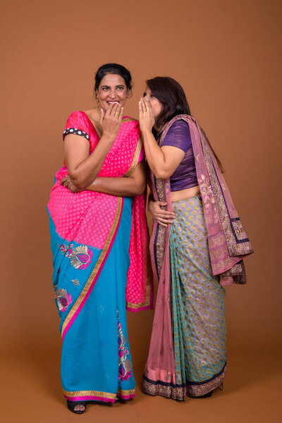 Studio shot of two mature Indian women wearing Sari Indian traditional clothes together against brown background