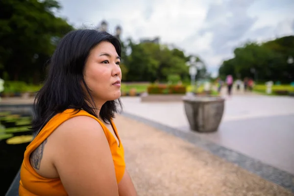 Profile view of overweight Asian woman sitting and thinking in park — Stock Photo, Image