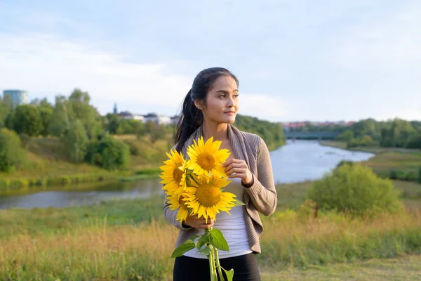 Jonge mooie Aziatische vrouw met boeket van zonnebloemen denken tegen ontspannen uitzicht op natuur — Stockfoto