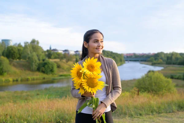 Jonge gelukkig Aziatische vrouw bedrijf boeket van zonnebloemen tegen ontspannen uitzicht op natuur — Stockfoto