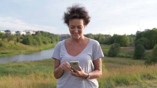 Feliz madura bela mulher pensando ao usar o telefone contra a visão relaxante da natureza — Vídeo de Stock