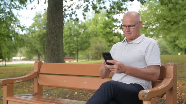 Šťastný starší muž sedí a používání telefonu v parku — Stock video