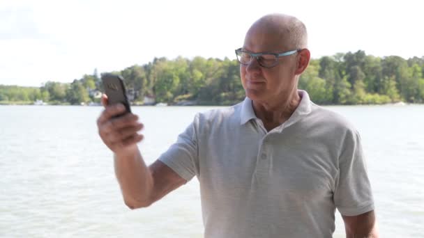 Feliz hombre mayor tomando selfie con el teléfono junto al lago — Vídeo de stock