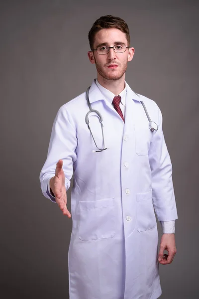 Junger schöner Mann Arzt trägt Brille gegen grauen Backgr — Stockfoto