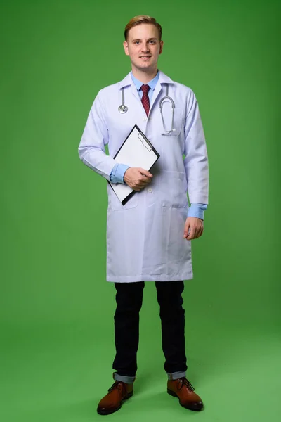 Young handsome man doctor with blond hair against green backgrou — Stock Photo, Image