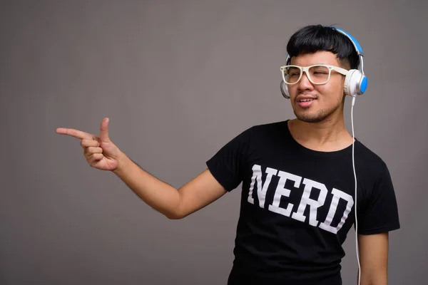 Young Asian nerd man listening to music against gray background — Stock Photo, Image