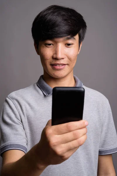 Молодий азіат користується мобільним телефоном проти сірого фону. — стокове фото