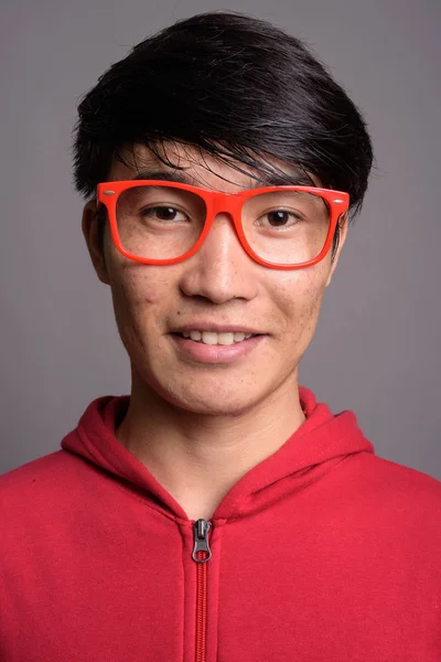 Junger asiatischer Mann mit roter Jacke und Brille gegen Grau — Stockfoto