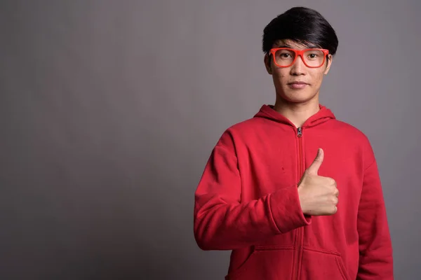 Młoda Azji man noszenia czerwonej kurtce z okularów przeciwko szary — Zdjęcie stockowe