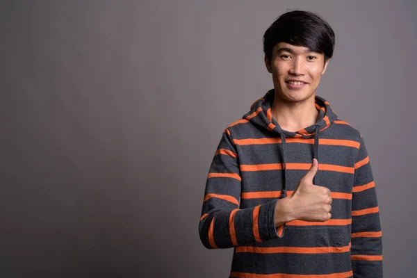 Young Asian man wearing striped hoodie against gray background — Stock Photo, Image