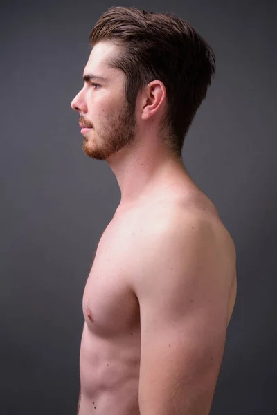 Mladý pohledný vousatý muž bez košile proti šedému pozadí — Stock fotografie