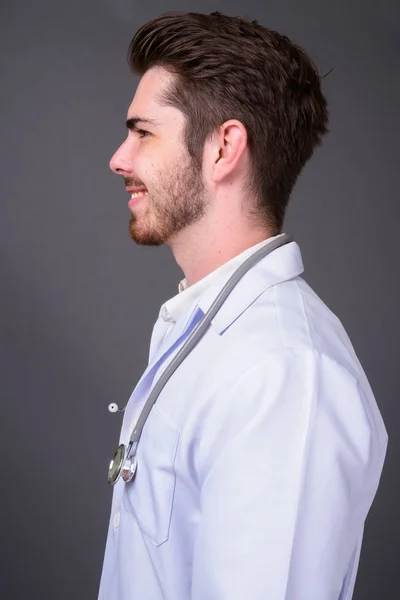 Junger gutaussehender bärtiger Mann Arzt vor grauem Hintergrund — Stockfoto