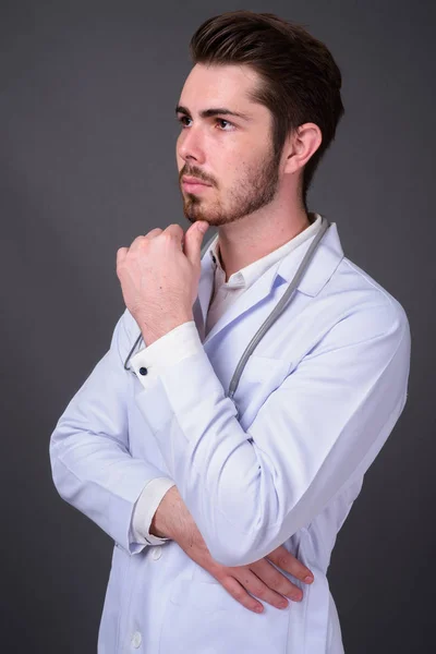 Junger gutaussehender bärtiger Mann Arzt vor grauem Hintergrund — Stockfoto