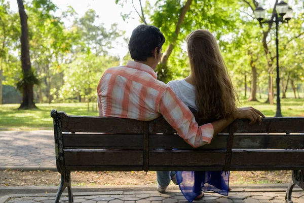 Visão traseira do casal multi étnico sentado no banco de madeira no amor — Fotografia de Stock