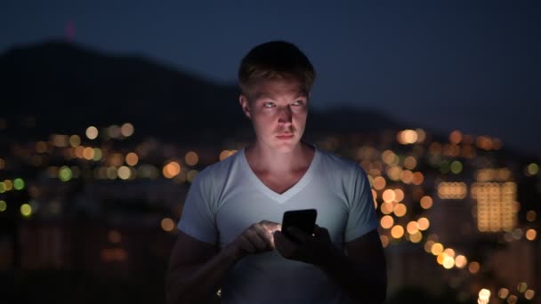 Jonge gelukkig toeristische Man met behulp van telefoon tegen de weergave van de stadslichten nachts — Stockvideo