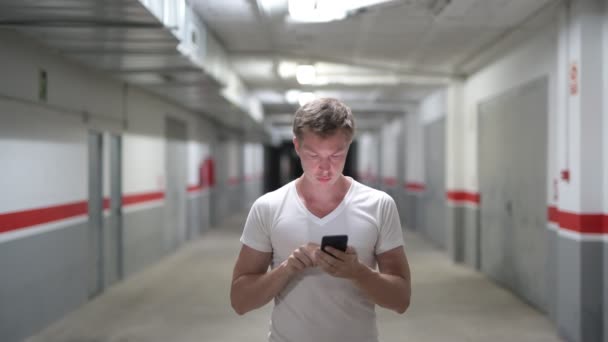 Счастливый молодой человек, использующий телефон вдоль коридора — стоковое видео