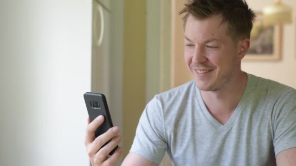 Joven hombre feliz usando el teléfono por la ventana — Vídeo de stock