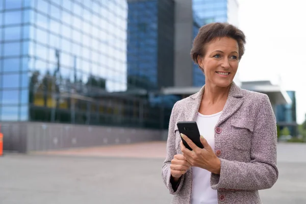 Счастливая пожилая деловая женщина думает, используя телефон на открытом воздухе — стоковое фото