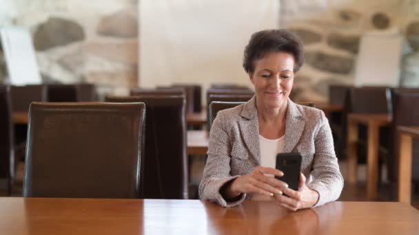 Щаслива зріла бізнес-леді думає під час використання телефону в кав'ярні — стокове відео