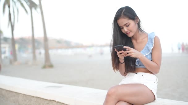 Счастливая азиатская женщина сидит с телефоном на городском пляже — стоковое видео