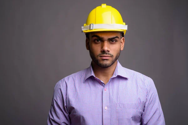 Young handsome Indian businessman wearing hardhat against gray b