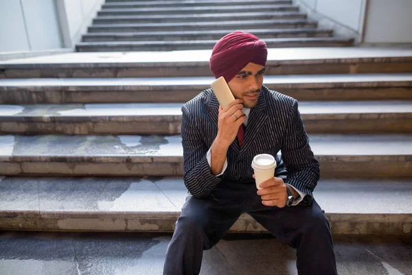 Homme d'affaires indien assis sur les escaliers tout en utilisant le téléphone et en buvant du café — Photo