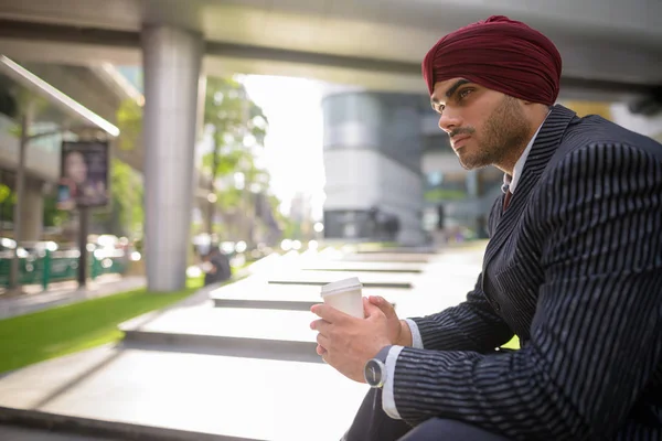 Indian businessman sitting outdoors in city while holding coffee cup