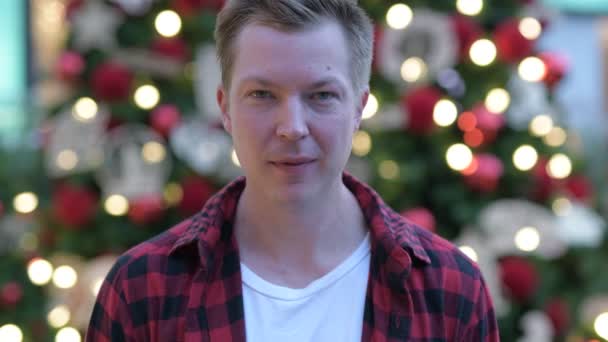 Šťastný mladý pohledný muž proti osvětlené vánoční stromy venku — Stock video