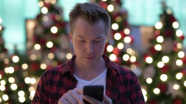 Jovem feliz sorrindo ao usar o telefone contra árvores de Natal ao ar livre — Vídeo de Stock