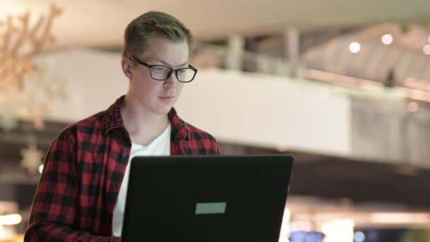Молодой симпатичный хипстер улыбается во время использования ноутбука в городе ночью — стоковое видео