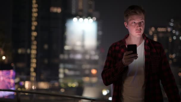 Jovem bonitão usando telefone contra vista da cidade à noite — Vídeo de Stock