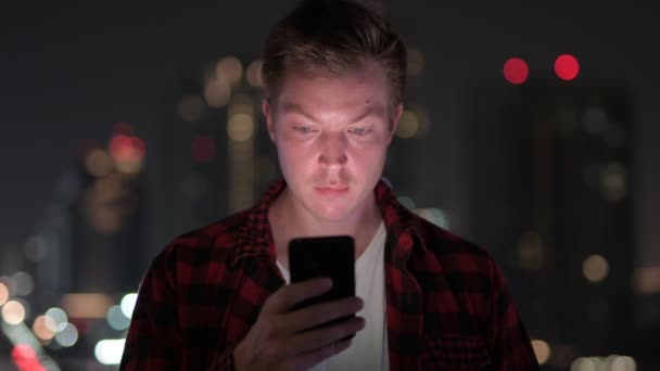 Молодой симпатичный хипстер разговаривает по телефону с видом на город ночью — стоковое видео