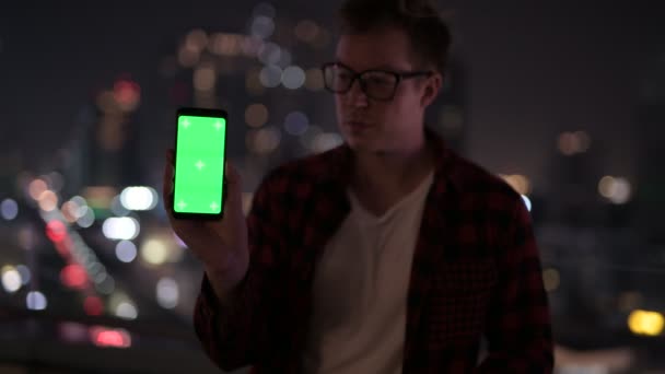 Fiatal, boldog csípő ember mutatja telefon ellen kilátás nyílik a városra, az éjszakai — Stock videók