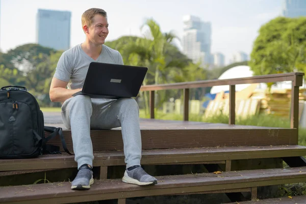 Χαρούμενος νεαρός σκέψης κατά τη χρήση του laptop σε ξύλινη γέφυρα στο πάρκο — Φωτογραφία Αρχείου