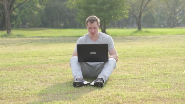 Joven hombre guapo sentado mientras usa el portátil en el parque — Vídeos de Stock