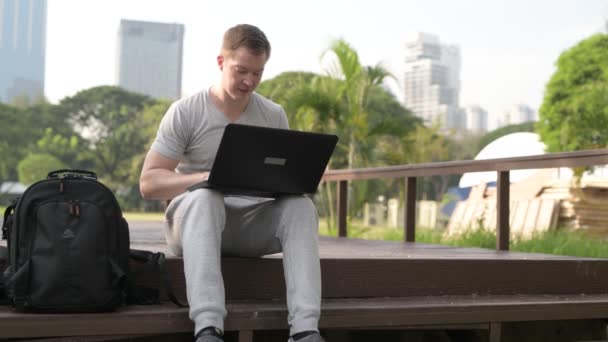 Joven feliz sentado en el puente de madera mientras usa el portátil en el parque — Vídeos de Stock