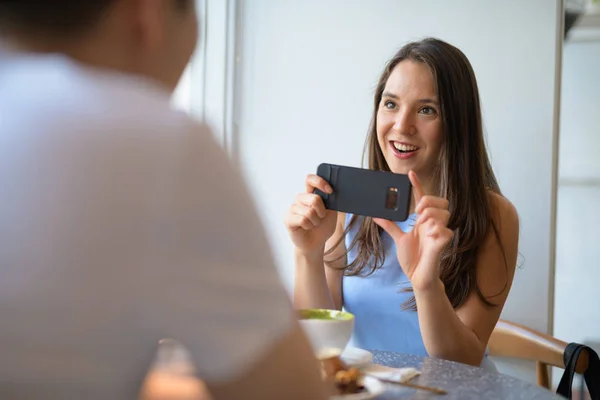 Joven mujer feliz tomando fotos de hombre joven con teléfono en la cafetería — Foto de Stock