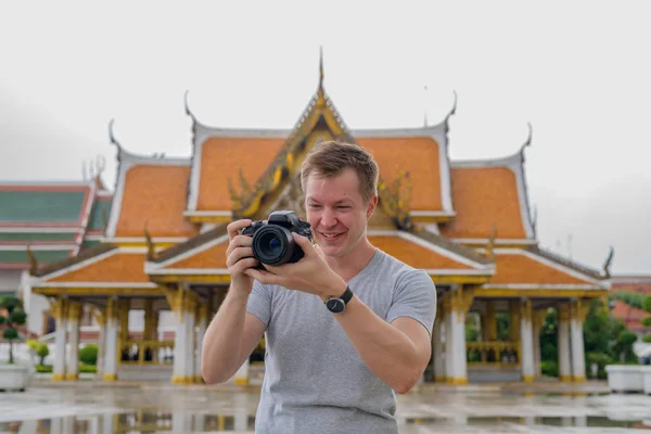 Young happy tourist man taking picture with camera against view of the Buddhist temple in Bangkok