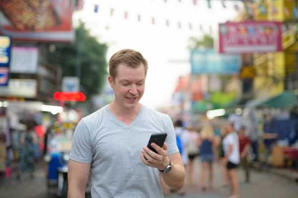 Young happy tourist man smiling while using phone against view of Khao San road in Bangkok — Stock Photo, Image