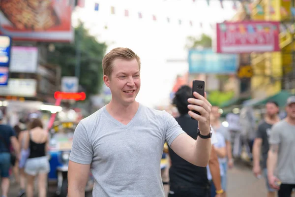 Junger glücklicher Touristenmann macht Selfie mit Blick auf belebte Straße in Khao san in Bangkok — Stockfoto