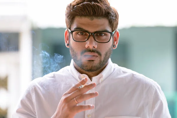 stock image Face of young bearded Indian man with eyeglasses smoking outdoors