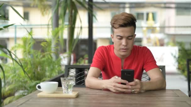 Gelukkig jonge Aziatische man met behulp van de telefoon bij de koffieshop buitenshuis — Stockvideo