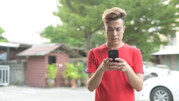 Szczęśliwy młody człowiek azjatyckiej uśmiechający się podczas korzystania z telefonu na zewnątrz — Wideo stockowe