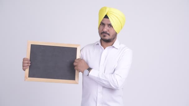 Benadrukt bebaarde Indische Sikh zakenman holding blackboard en duimen neer geven — Stockvideo